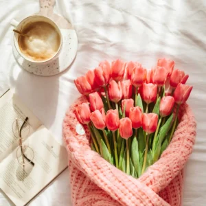 Flatlay of coral pink tulips wrapped in a knit sweater, next to a cup of coffee, open book, and glasses on a white bed. A peaceful visual for Morning Prayer for Today.