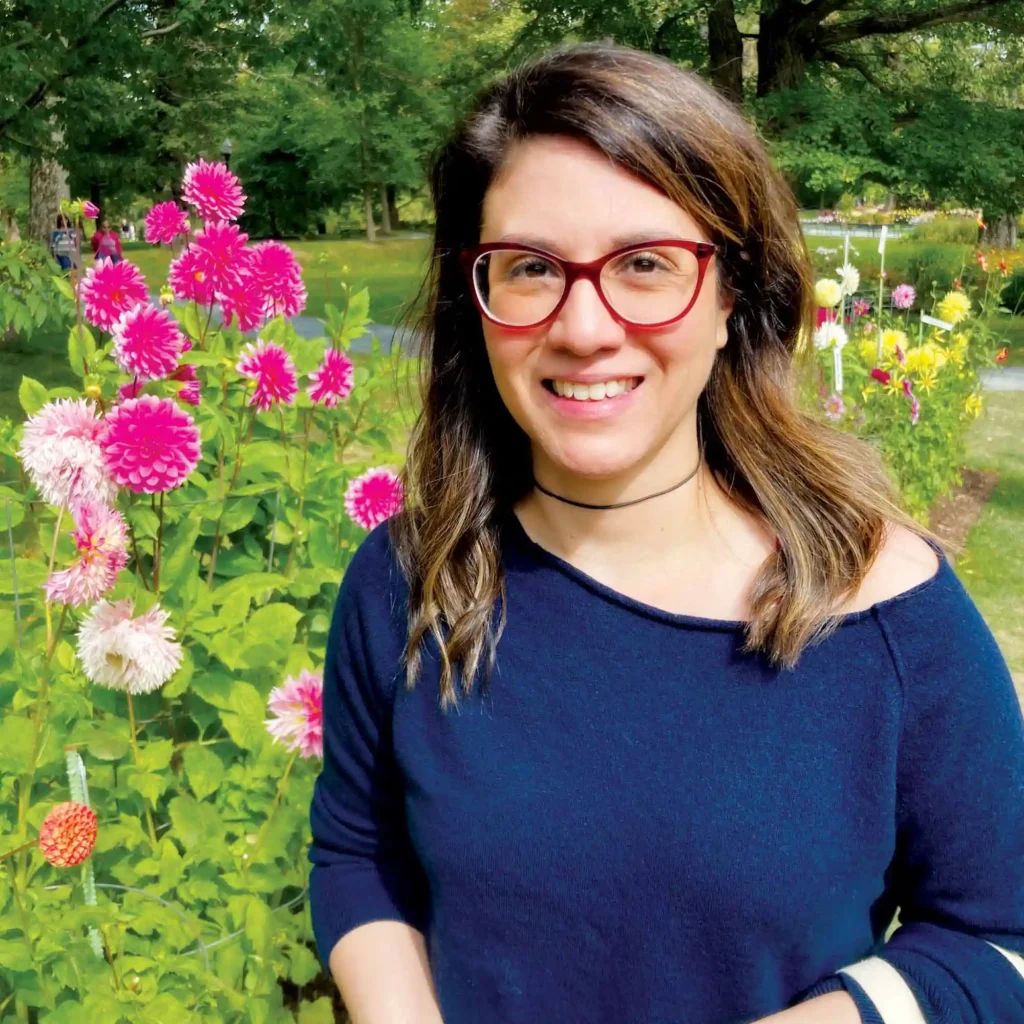 A smiling Woman with red glasses and a blue sweater standing next to pink flowers